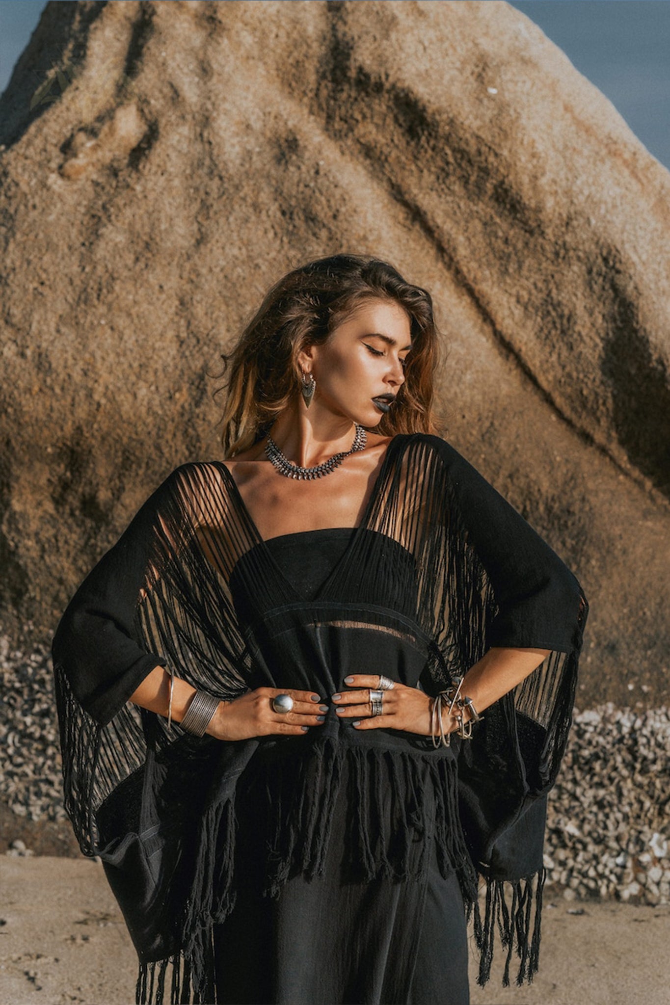 A woman stands confidently on a beach with a large rock in the background. She is wearing a black fringe shawl and an array of jewelry, including necklaces, rings, and bracelets. The sunlight casts dramatic shadows on her Hand Loomed Cotton Blouse from AYA Sacred Wear, highlighting its distinctive boho style.