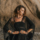 A woman stands confidently on a beach with a large rock in the background. She is wearing a black fringe shawl and an array of jewelry, including necklaces, rings, and bracelets. The sunlight casts dramatic shadows on her Hand Loomed Cotton Blouse from AYA Sacred Wear, highlighting its distinctive boho style.