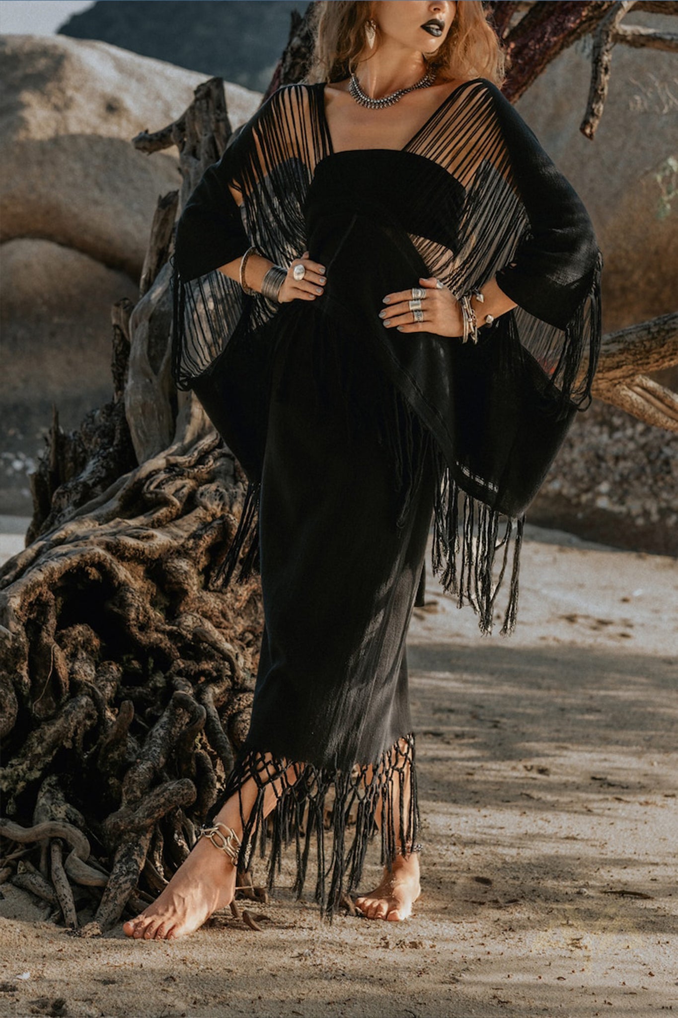 Standing on a sandy beach near driftwood, a person is adorned in a black dress featuring a fringed shawl and hemline. Their look is accentuated by the Hand Loomed Cotton Blouse from AYA Sacred Wear, harmonizing with an array of rings, necklaces, and bracelets to complement their boho style. In the background lies a rugged rocky landscape.