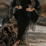 Standing on a sandy beach near driftwood, a person is adorned in a black dress featuring a fringed shawl and hemline. Their look is accentuated by the Hand Loomed Cotton Blouse from AYA Sacred Wear, harmonizing with an array of rings, necklaces, and bracelets to complement their boho style. In the background lies a rugged rocky landscape.