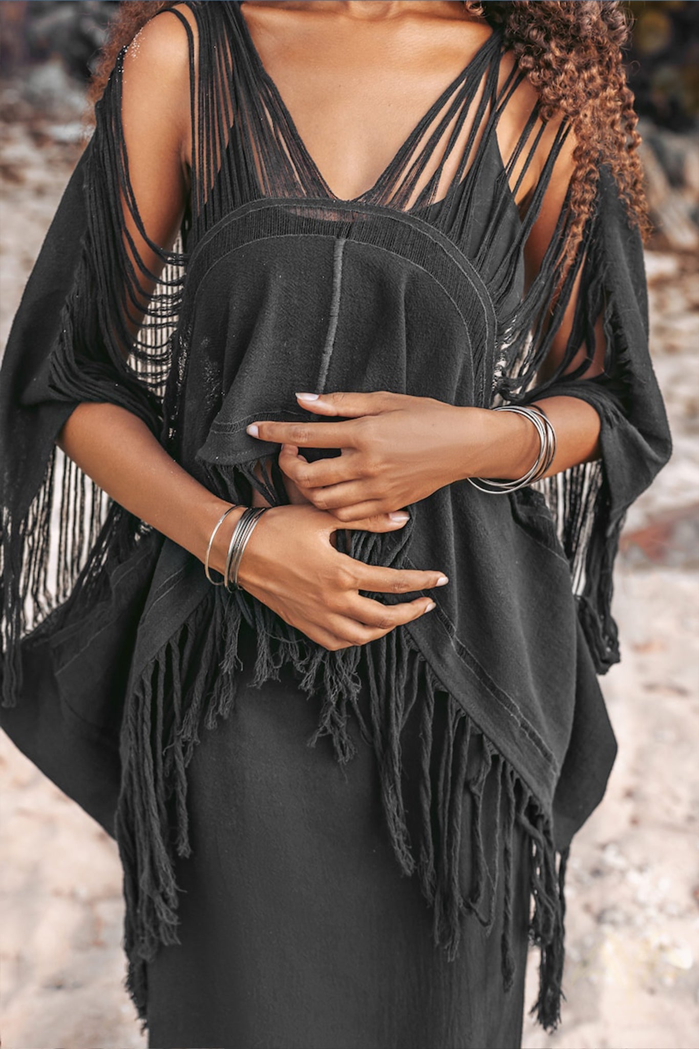 A person wearing a bohemian top from AYA Sacred Wear stands outdoors, exuding a boho vibe. Their hands are crossed in front of them, and their curly hair cascades over their shoulders. The blurred natural background complements the look beautifully.
