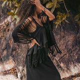 Wearing the Hand Loomed Cotton Blouse from AYA Sacred Wear, a woman poses effortlessly on sandy ground amidst verdant leaves and a rocky backdrop. Her arm is gracefully arched with her hand resting against her head, showcasing the timeless style of this boho top.