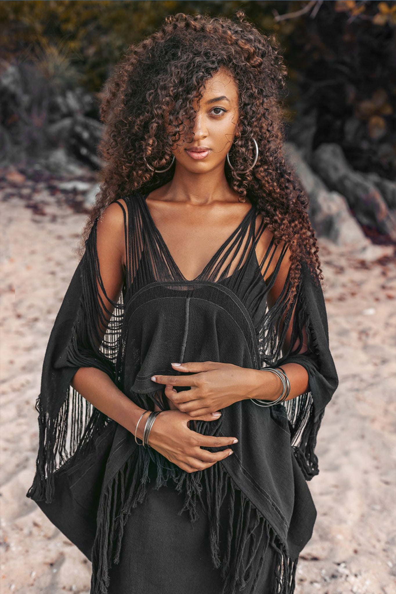 A woman with curly hair stands on a sandy beach, wearing an AYA Sacred Wear Hand Loomed Cotton Blouse Bohemian Top and hoop earrings. She looks at the camera with her hands gently clasped in front. Trees are visible in the background.