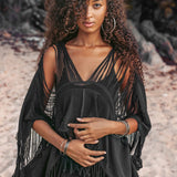 A woman with curly hair stands on a sandy beach, wearing an AYA Sacred Wear Hand Loomed Cotton Blouse Bohemian Top and hoop earrings. She looks at the camera with her hands gently clasped in front. Trees are visible in the background.