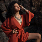 A woman poses confidently in a handmade, knotted, red mini organic jumpsuit, showcasing its flowing sleeves and cinched waist. She accessorizes with a long necklace and statement rings, set against a textured rock background that adds depth to the scene. The warm tones of the outfit contrast beautifully with the natural surroundings, highlighting the jumpsuit’s bohemian style.