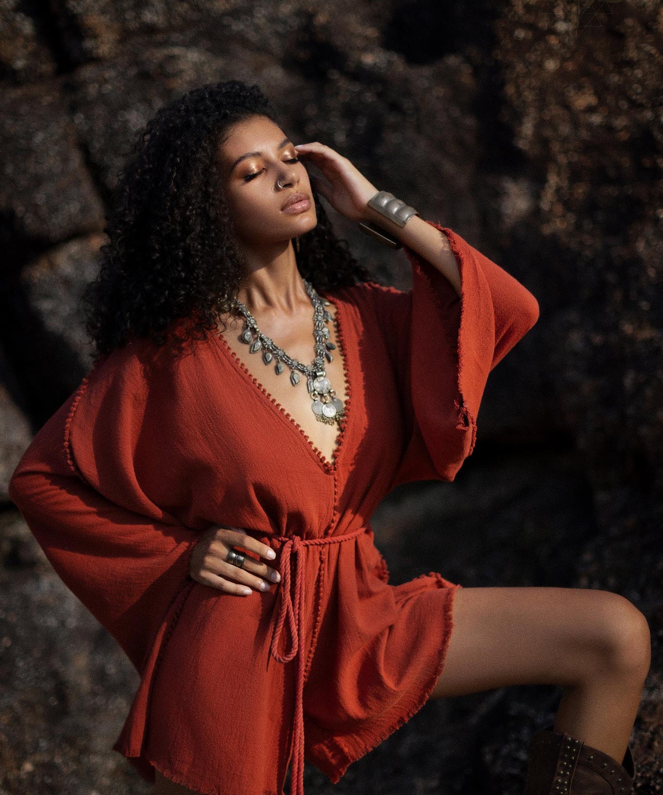A woman poses confidently in a handmade, knotted, red mini organic jumpsuit, showcasing its flowing sleeves and cinched waist. She accessorizes with a long necklace and statement rings, set against a textured rock background that adds depth to the scene. The warm tones of the outfit contrast beautifully with the natural surroundings, highlighting the jumpsuit’s bohemian style.