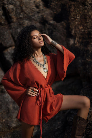 A woman poses confidently in a handmade, knotted, red mini organic jumpsuit, showcasing its flowing sleeves and cinched waist. She accessorizes with a long necklace and statement rings, set against a textured rock background that adds depth to the scene. The warm tones of the outfit contrast beautifully with the natural surroundings, highlighting the jumpsuit’s bohemian style.