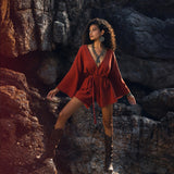 A woman poses on rocky terrain, wearing a handmade knotted red mini jumpsuit. The outfit features long sleeves and a tie waist, complemented by tall brown boots and statement jewelry. The natural setting enhances the organic vibe of the romper dress.