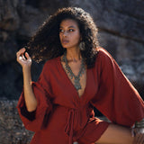 A woman poses confidently in a handmade, knotted red mini organic jumpsuit, showcasing a deep V-neck and flowing bell sleeves. She accessorizes with bold statement jewelry, including large earrings and a layered necklace. The backdrop features natural rock formations, enhancing the bohemian vibe of the outfit. Perfect for a stylish summer look, this romper dress combines comfort and chicness.