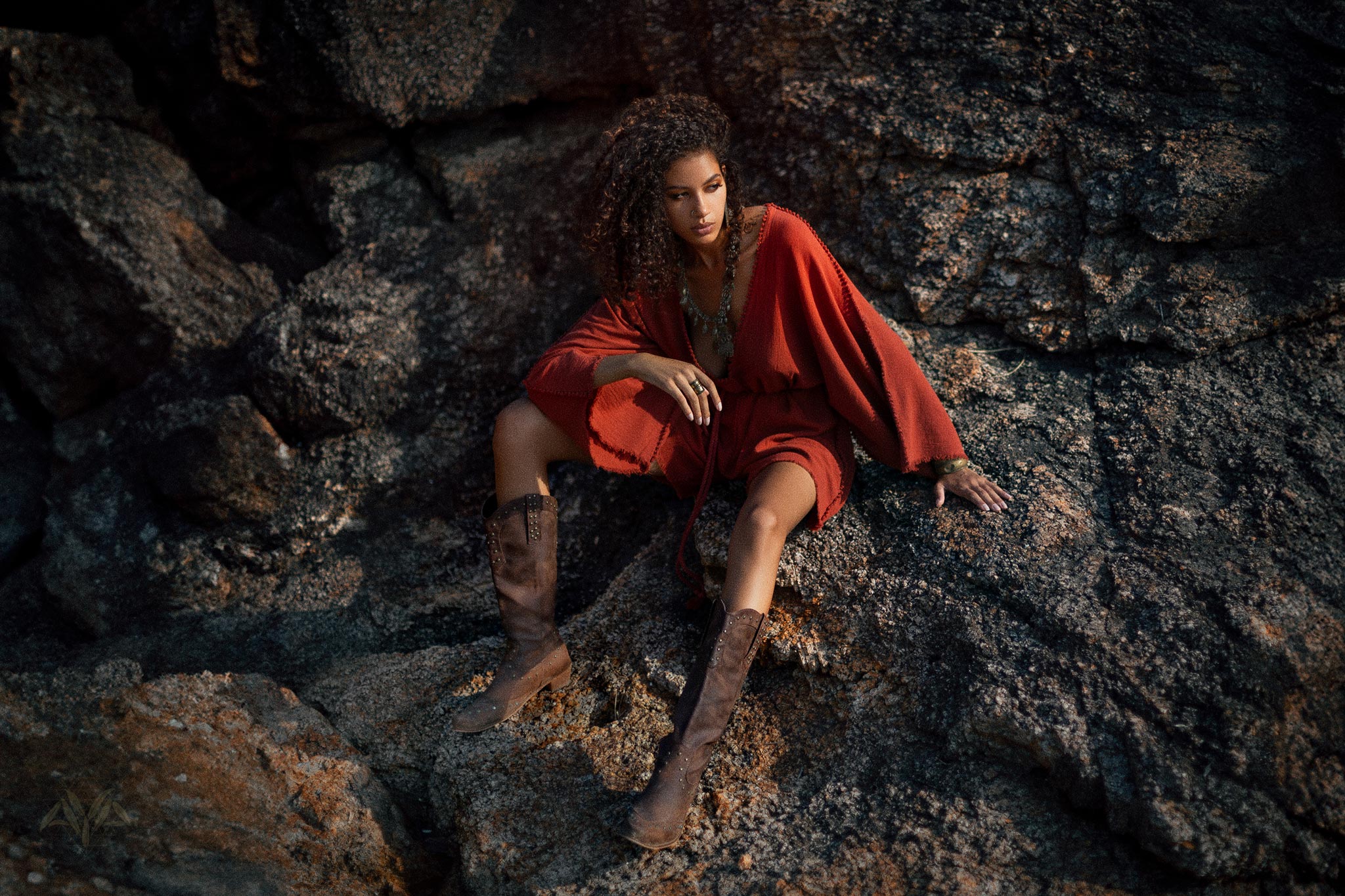 A woman sits confidently on rocky terrain, wearing a handmade knotted red mini organic jumpsuit. The jumpsuit features a relaxed fit and wide sleeves, complementing her stylish brown knee-high boots. Her tousled hair frames her face, and bold accessories add a touch of elegance to the casual look. The natural setting highlights the earthy tones of her outfit.