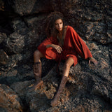 A woman sits confidently on rocky terrain, wearing a handmade knotted red mini organic jumpsuit. The jumpsuit features a relaxed fit and wide sleeves, complementing her stylish brown knee-high boots. Her tousled hair frames her face, and bold accessories add a touch of elegance to the casual look. The natural setting highlights the earthy tones of her outfit.
