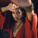 A woman with curly hair poses in a striking red handmade knotted jumpsuit. The jumpsuit features a deep V-neck and flowing sleeves that seem to catch the light. She is adorned with a bold silver necklace and oversized earrings, and her expression is confident and captivating, as she shields her eyes from the sun. This image embodies the essence of a stylish organic romper dress for women, perfect for any casual outing.