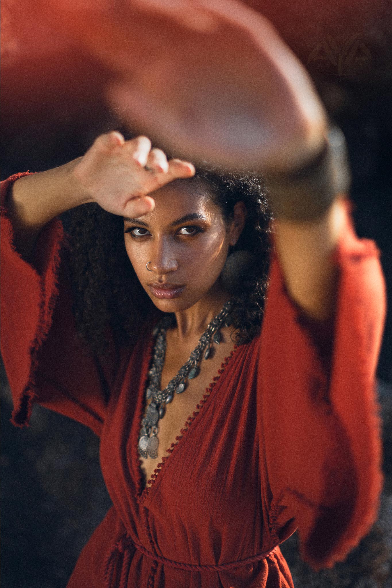 A woman with curly hair poses in a striking red handmade knotted jumpsuit. The jumpsuit features a deep V-neck and flowing sleeves that seem to catch the light. She is adorned with a bold silver necklace and oversized earrings, and her expression is confident and captivating, as she shields her eyes from the sun. This image embodies the essence of a stylish organic romper dress for women, perfect for any casual outing.