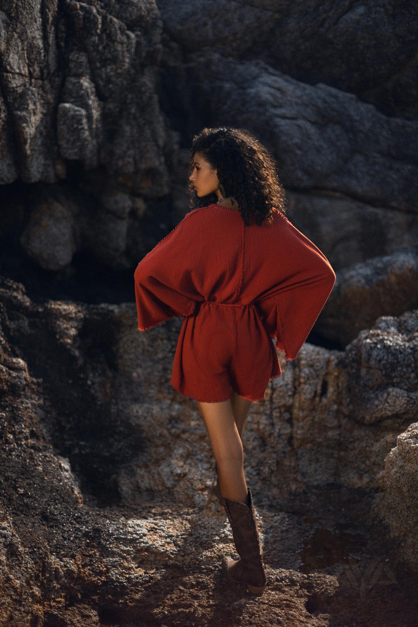 A woman stands facing away, showcasing a handmade knotted red mini organic jumpsuit. The romper features wide sleeves and a cinched waist, complemented by stylish brown boots. The background features rocky textures, contributing to a natural outdoor setting.