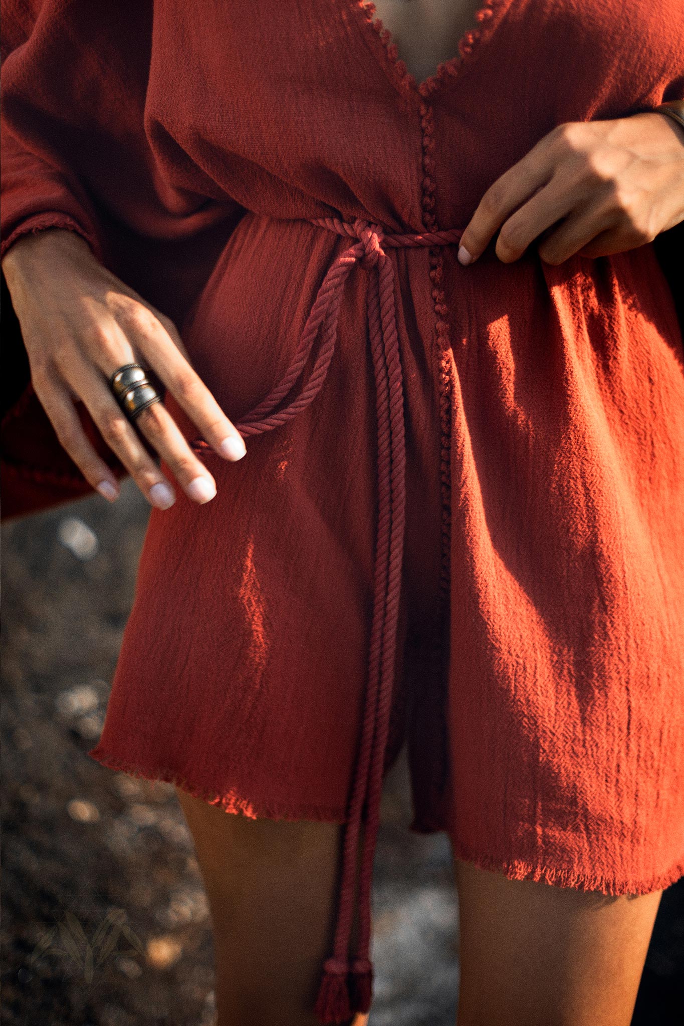A close-up of a person holding the tie of a Handmade Knotted Jumpsuit in red. The garment features a v-neckline, buttons down the front, and a cinched waist with a braided belt. The fabric appears soft and organic, emphasizing the casual yet stylish nature of this mini romper dress for women.