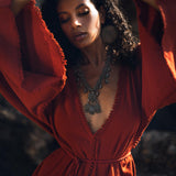 A woman gracefully poses in a Handmade Knotted Red Mini Organic Jumpsuit Romper Dress. The jumpsuit features flowing sleeves and a deep V-neckline, accentuated by a statement silver necklace and large earrings. The background is softly blurred, highlighting the elegant style of the outfit.