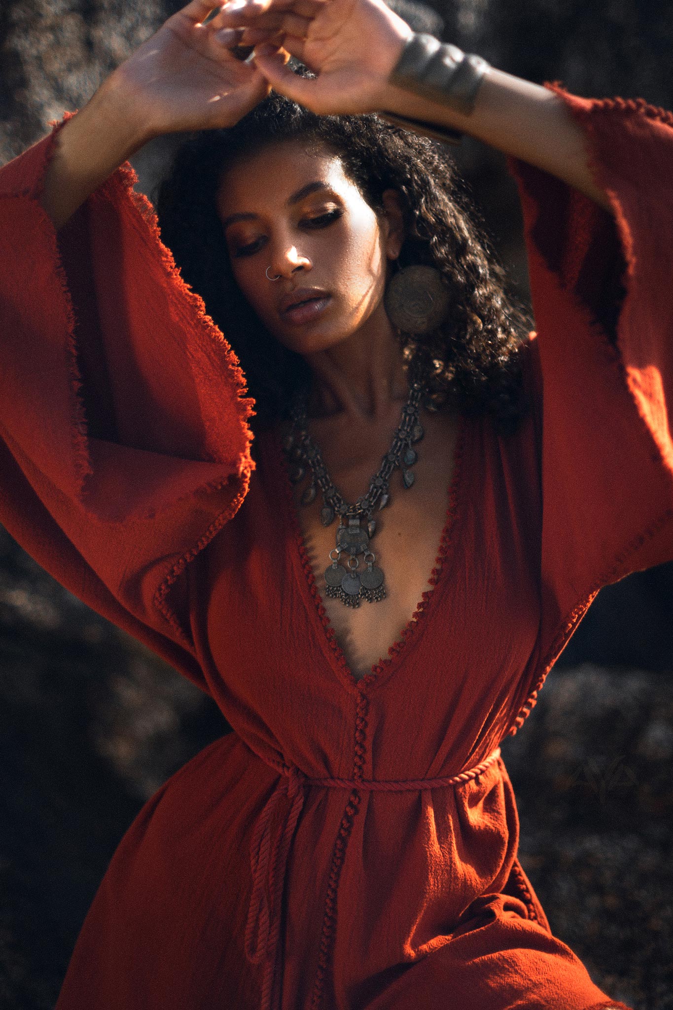 A woman gracefully poses in a Handmade Knotted Red Mini Organic Jumpsuit Romper Dress. The jumpsuit features flowing sleeves and a deep V-neckline, accentuated by a statement silver necklace and large earrings. The background is softly blurred, highlighting the elegant style of the outfit.