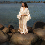 A woman in a flowing Cream Silk Kimono Cover Up by AYA Sacred Wear stands barefoot on large rocks near a tranquil body of water. Her gaze is directed left, and lush green hills rise in the background under a clear sky.