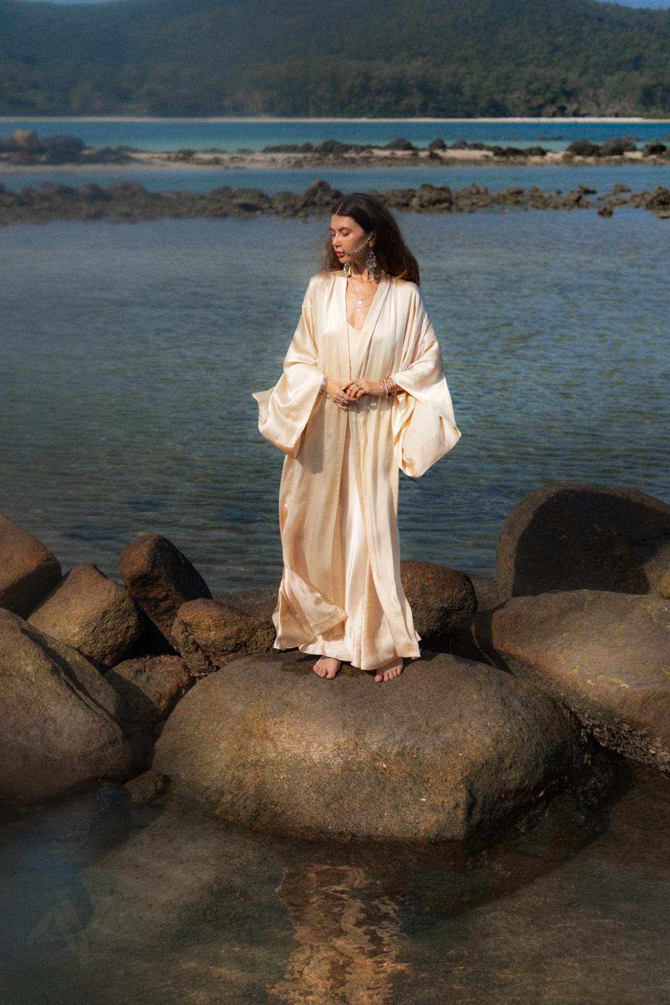 A woman in a flowing Cream Silk Kimono Cover Up by AYA Sacred Wear stands barefoot on large rocks near a tranquil body of water. Her gaze is directed left, and lush green hills rise in the background under a clear sky.