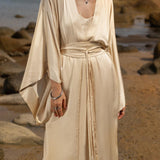 On a rocky beach, someone wears the AYA Sacred Wear Cream Silk Kimono Cover Up, a wide sleeve bohemian cardigan. The deep V-neck and Ahimsa silk add elegance, enhanced by layered necklaces and statement earrings. The sea backdrop embodies sustainable living perfectly.