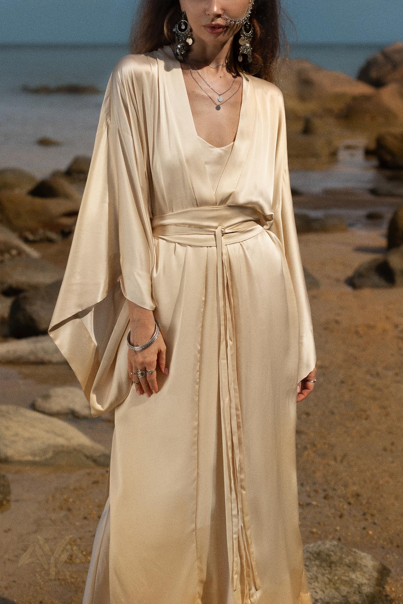 A person stands on a rocky beach, draped in an elegant Cream Silk Kimono Cover Up by AYA Sacred Wear, exuding the luxurious essence of fine silk kimonos. This bohemian cardigan features wide sleeves and a deep V-neckline, complete with long waist ties for added style. Layered necklaces and large dangling earrings complement their chic ensemble.