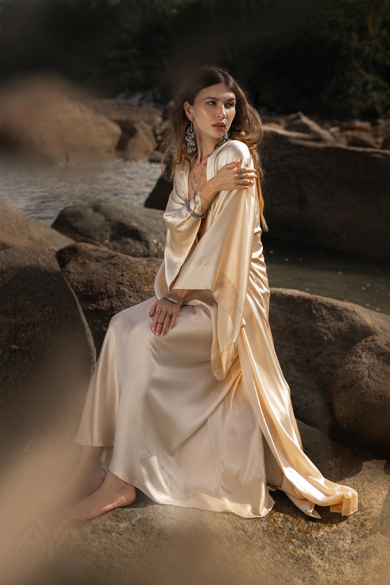 A woman in the NEW! Cream Silk Kimono Cover Up by AYA Sacred Wear, featuring wide sleeves and a bohemian cardigan style, sits on large rocks by the water, adorned with striking silver jewelry. Her hair cascades freely as she gazes thoughtfully into the distance. The scene captures a harmonious blend of calm and natural beauty, reflecting principles of sustainable living.