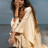 A woman with long hair sits on a rock by the water, dressed in a flowing Cream Silk Kimono Cover Up by AYA Sacred Wear. Her left hand covers part of her face adorned with various rings and bracelets, while the sky and sea provide a calm backdrop for sustainable living.