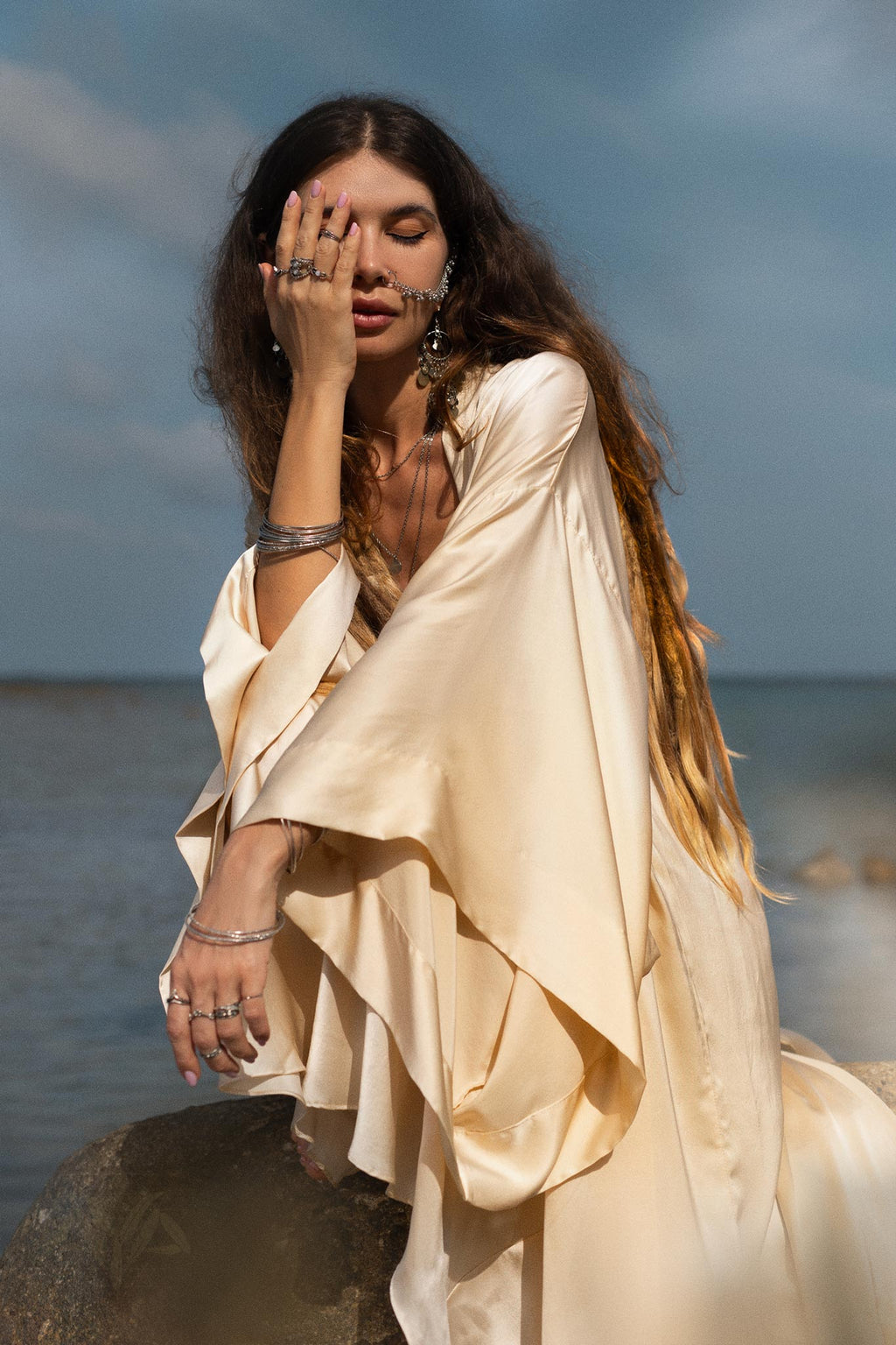 A woman with long hair sits on a rock by the water, dressed in a flowing Cream Silk Kimono Cover Up by AYA Sacred Wear. Her left hand covers part of her face adorned with various rings and bracelets, while the sky and sea provide a calm backdrop for sustainable living.