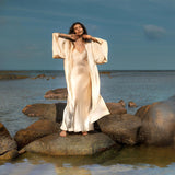A woman stands on rocks by the sea, wearing an AYA Sacred Wear Cream Silk Kimono Cover Up and a flowing dress. Her arms touch her head, embracing the calm sea and clear sky, reflecting a serene connection with nature and sustainable living.