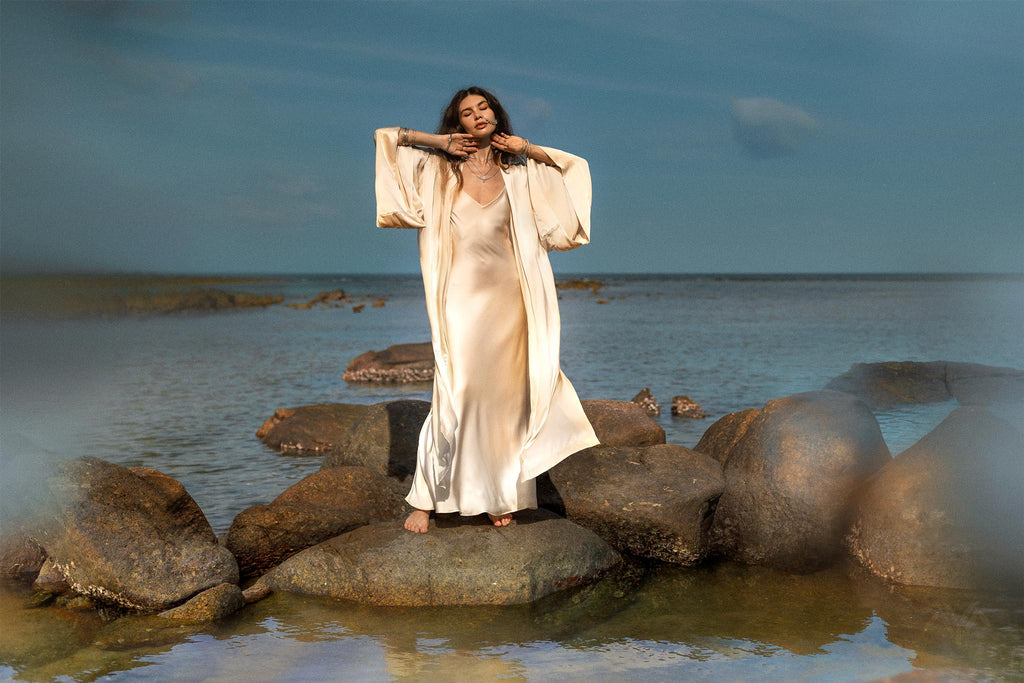 A woman wearing the NEW! Cream Silk Kimono Cover Up by AYA Sacred Wear stands on rocks near the sea. The wide-sleeved bohemian cardigan, made from Ahimsa silk, perfectly captures her graceful pose with hands near her face and eyes closed against a backdrop of clear skies and calm water—an embodiment of sustainable living.