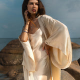 A woman poses contemplatively on seaside rocks, wearing a Cream Silk Kimono Cover Up with wide sleeves from AYA Sacred Wear. With one hand on her face and the other on her hip, she embraces sustainable living in a serene beach setting under clear skies and soft clouds.