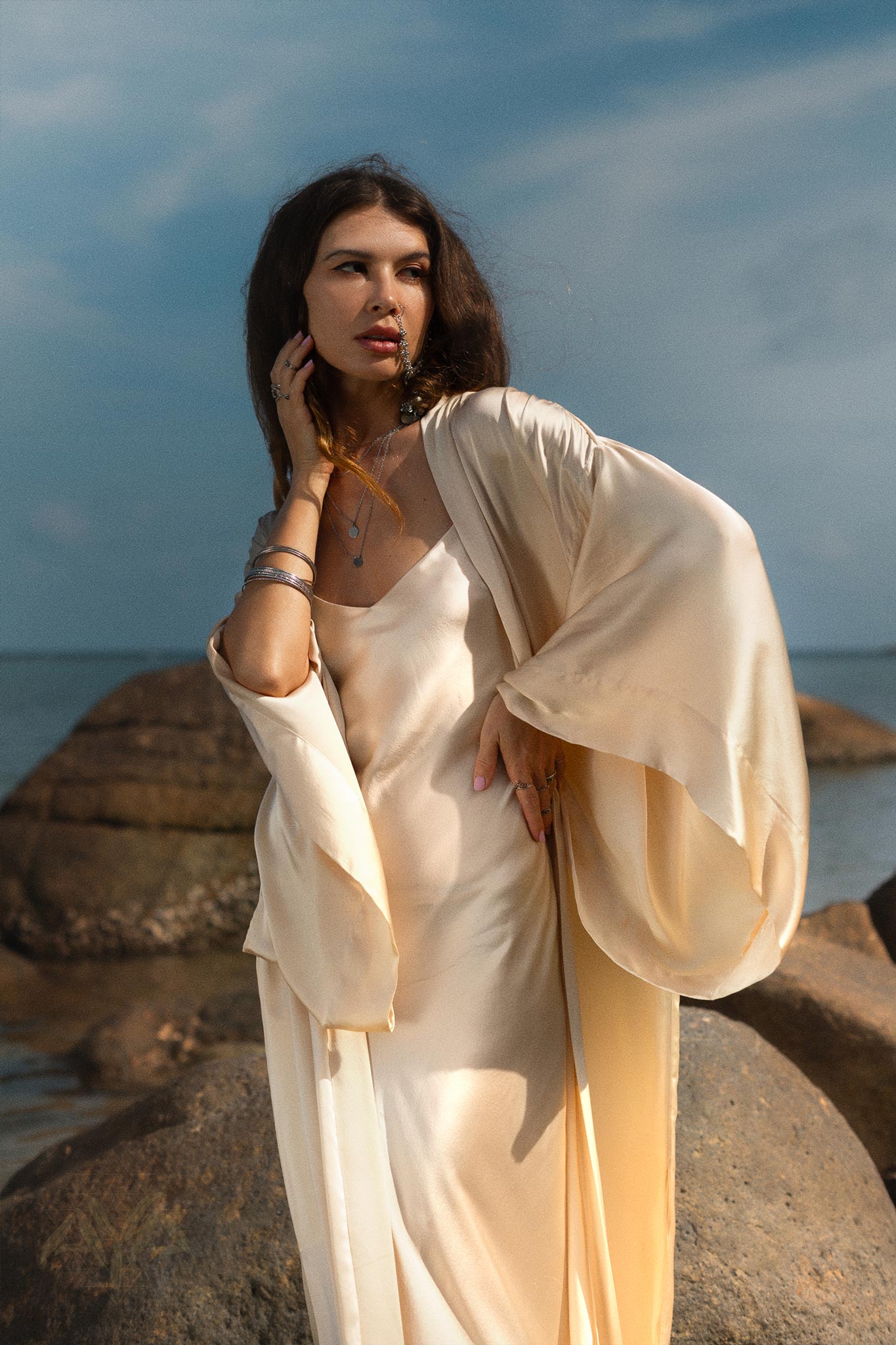 A woman poses contemplatively on seaside rocks, wearing a Cream Silk Kimono Cover Up with wide sleeves from AYA Sacred Wear. With one hand on her face and the other on her hip, she embraces sustainable living in a serene beach setting under clear skies and soft clouds.