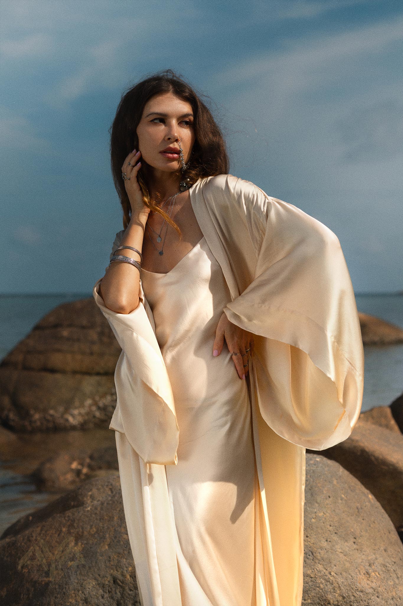 A woman stands on rocky terrain by the sea, draped elegantly in AYA Sacred Wear's NEW! Cream Silk Kimono Cover Up over a matching dress. She poses with one hand on her face and the other on her waist, gazing confidently into the distance. The sky is clear and blue, capturing a tranquil embrace of sustainable living.