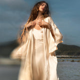 A woman stands near the water in a cream-colored AYA Sacred Wear Silk Kimono Cover Up with wide sleeves. She has long, wavy hair and wears several necklaces, her hands gently touching her chest. Behind her is a blurred landscape of hills and sky.