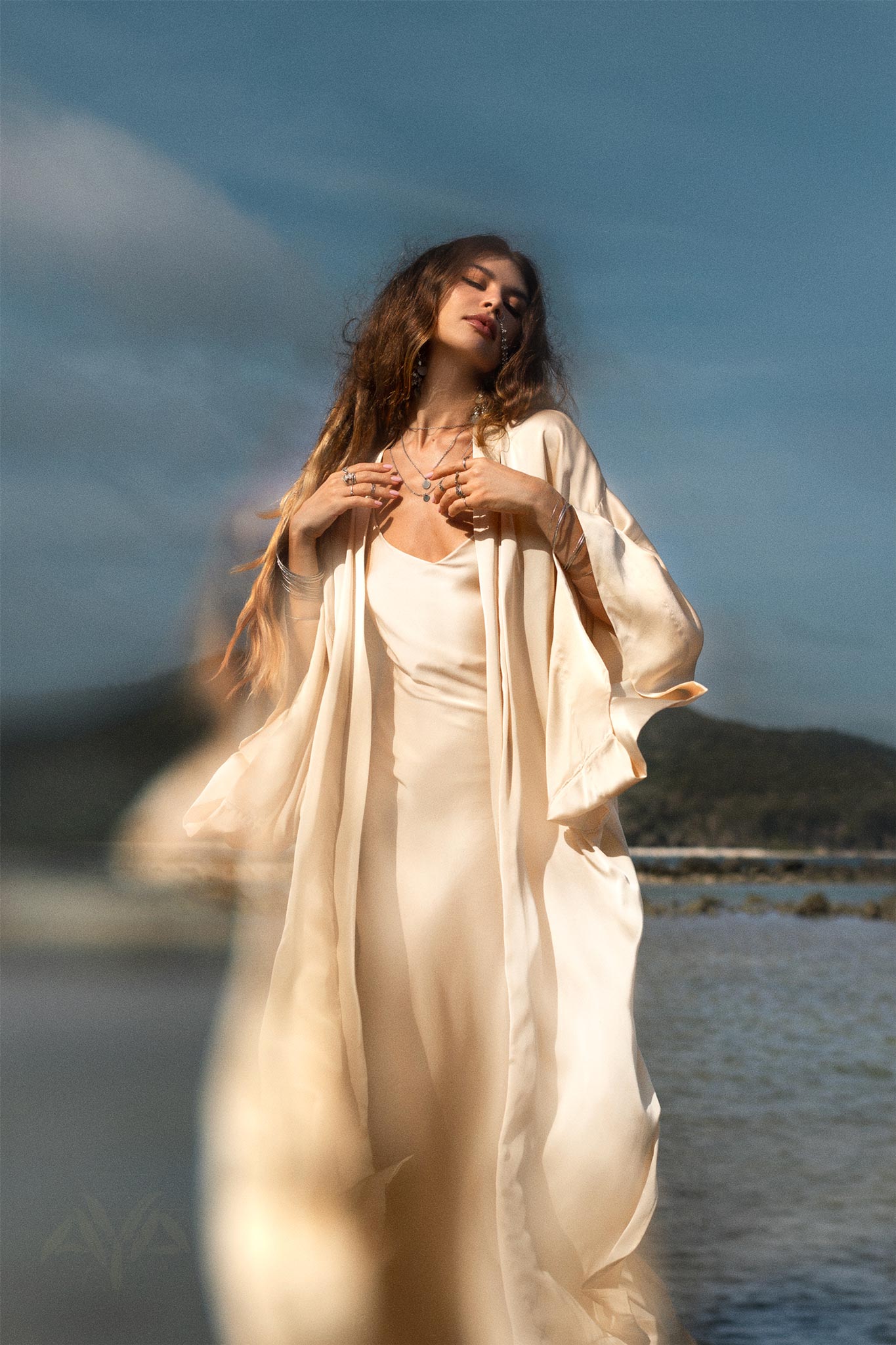 A woman stands near the water in a cream-colored AYA Sacred Wear Silk Kimono Cover Up with wide sleeves. She has long, wavy hair and wears several necklaces, her hands gently touching her chest. Behind her is a blurred landscape of hills and sky.
