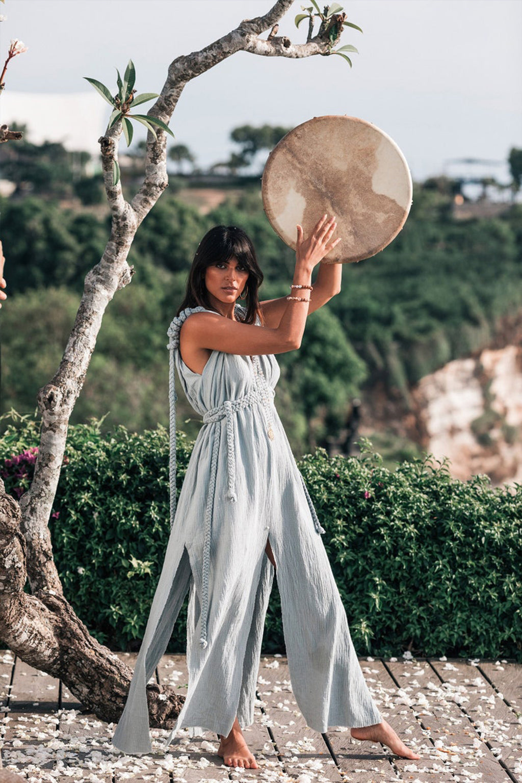 A woman wearing the Light Blue Nomad Spirit Dress from AYA Sacred Wear holds a large circular instrument above her head. She stands barefoot on a pebble-covered surface, surrounded by lush greenery and trees, with a picturesque landscape in the background.