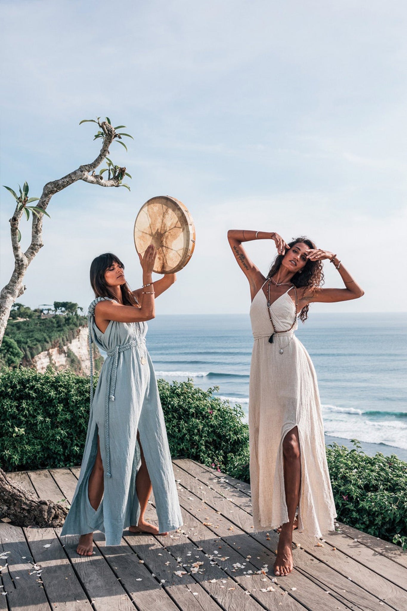 The setting features two women on a wooden deck with an ocean view. They both don flowing boho maxi dresses, one of them showcasing the Light Blue Nomad Spirit Dress from AYA Sacred Wear, designed in a plus size with adjustable features. As one woman plays a drum, the other strikes a playful pose. A tree and coastal scenery provide the backdrop under a clear sky.