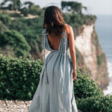A woman wearing the Light Blue Nomad Spirit Dress by AYA Sacred Wear stands on a wooden deck, facing away towards a scenic cliffside with lush greenery and the ocean below. Her long hair cascades down her back, and petals are scattered on the ground around her.