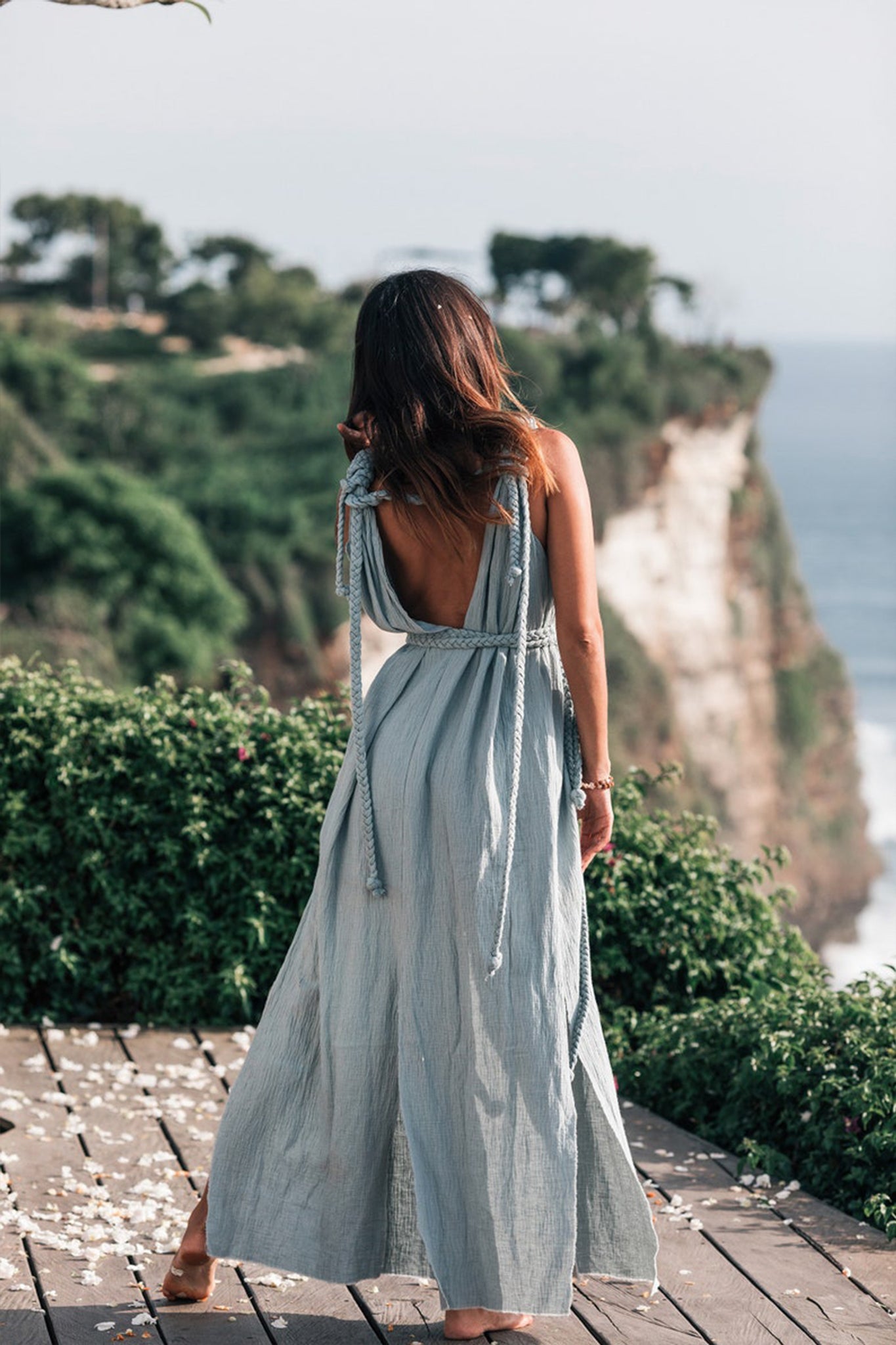 A woman wearing the Light Blue Nomad Spirit Dress by AYA Sacred Wear stands on a wooden deck, facing away towards a scenic cliffside with lush greenery and the ocean below. Her long hair cascades down her back, and petals are scattered on the ground around her.