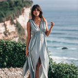 A woman wearing the AYA Sacred Wear Light Blue Nomad Spirit Dress, tailored for plus sizes with adjustable features, strolls barefoot on a wooden deck. She is near a cliffside with an ocean vista in the background, surrounded by lush greenery that enhances the tranquil coastal atmosphere.