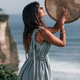 Outdoors by the seaside cliff, a woman stands holding a large round drum above her head, wearing the Light Blue Nomad Spirit Dress from AYA Sacred Wear. Her eyes are closed, surrounded by the serene ocean and vibrant greenery.