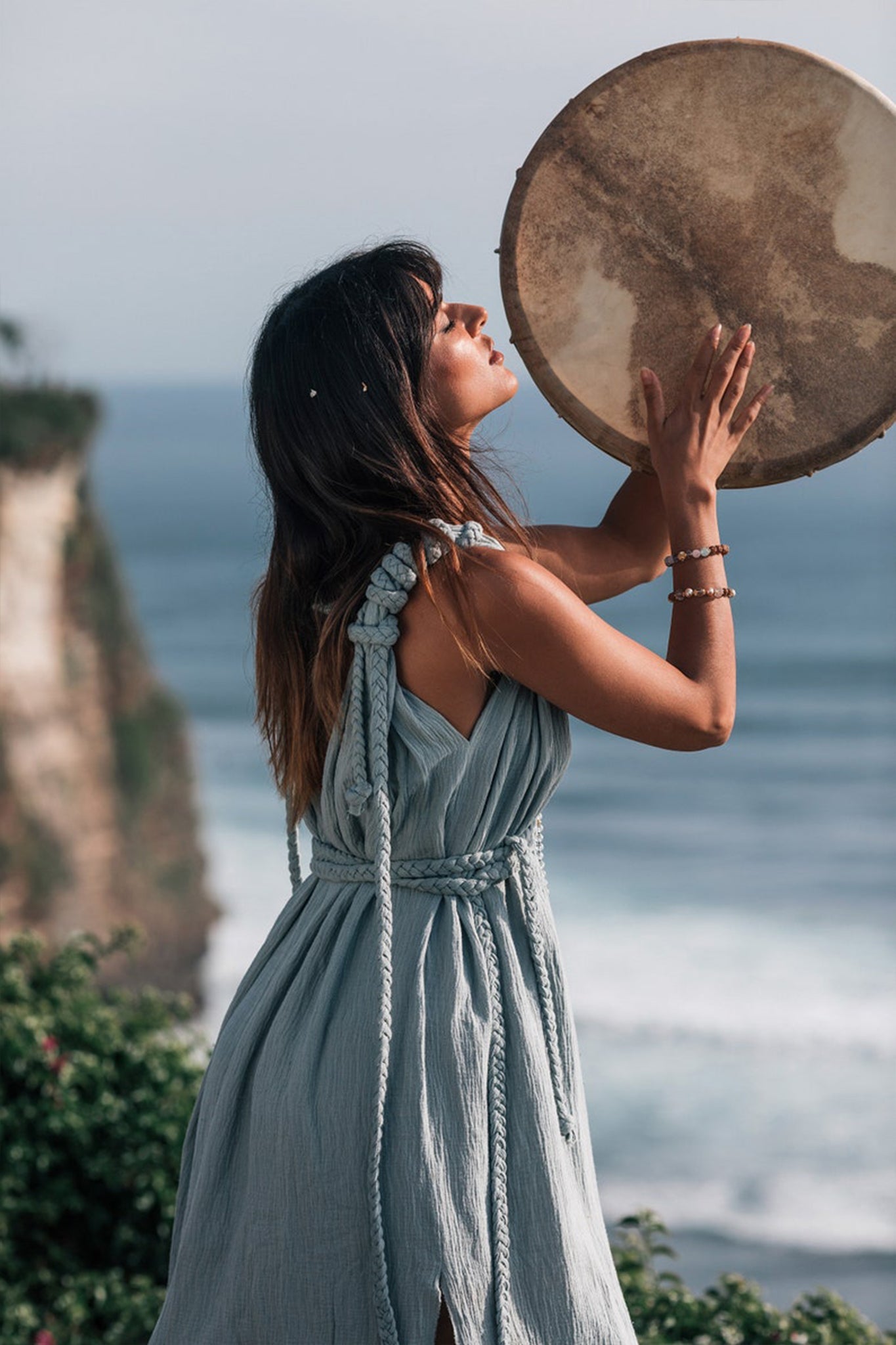 Outdoors by the seaside cliff, a woman stands holding a large round drum above her head, wearing the Light Blue Nomad Spirit Dress from AYA Sacred Wear. Her eyes are closed, surrounded by the serene ocean and vibrant greenery.