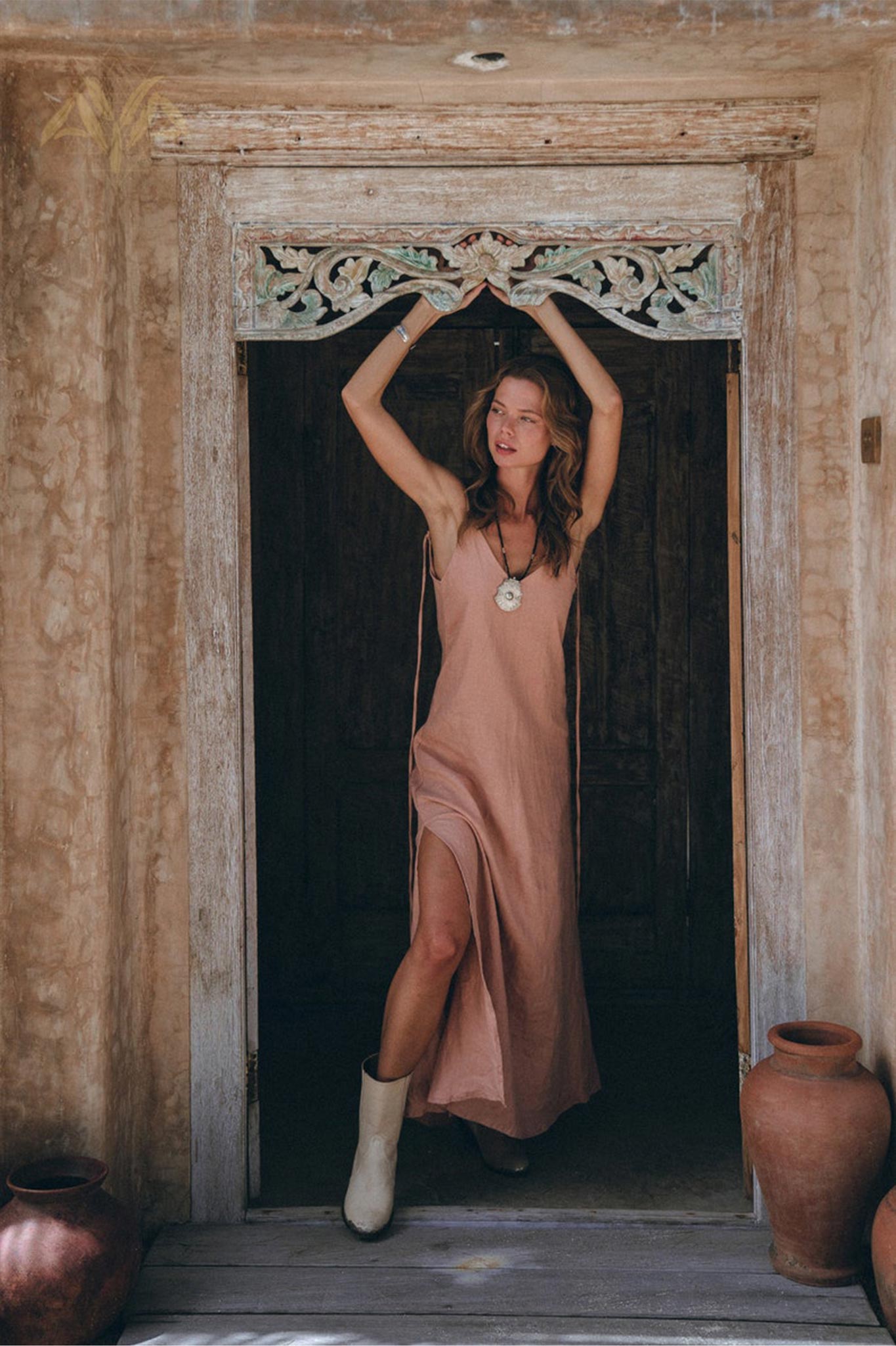 A woman in an elegant pink linen slip dress by AYA Sacred Wear stands in a rustic doorway, arms gracefully raised to touch the wooden frame. She complements her look with a pendant necklace and white boots, while two clay pots flank the entrance against a textured, earthy wall.