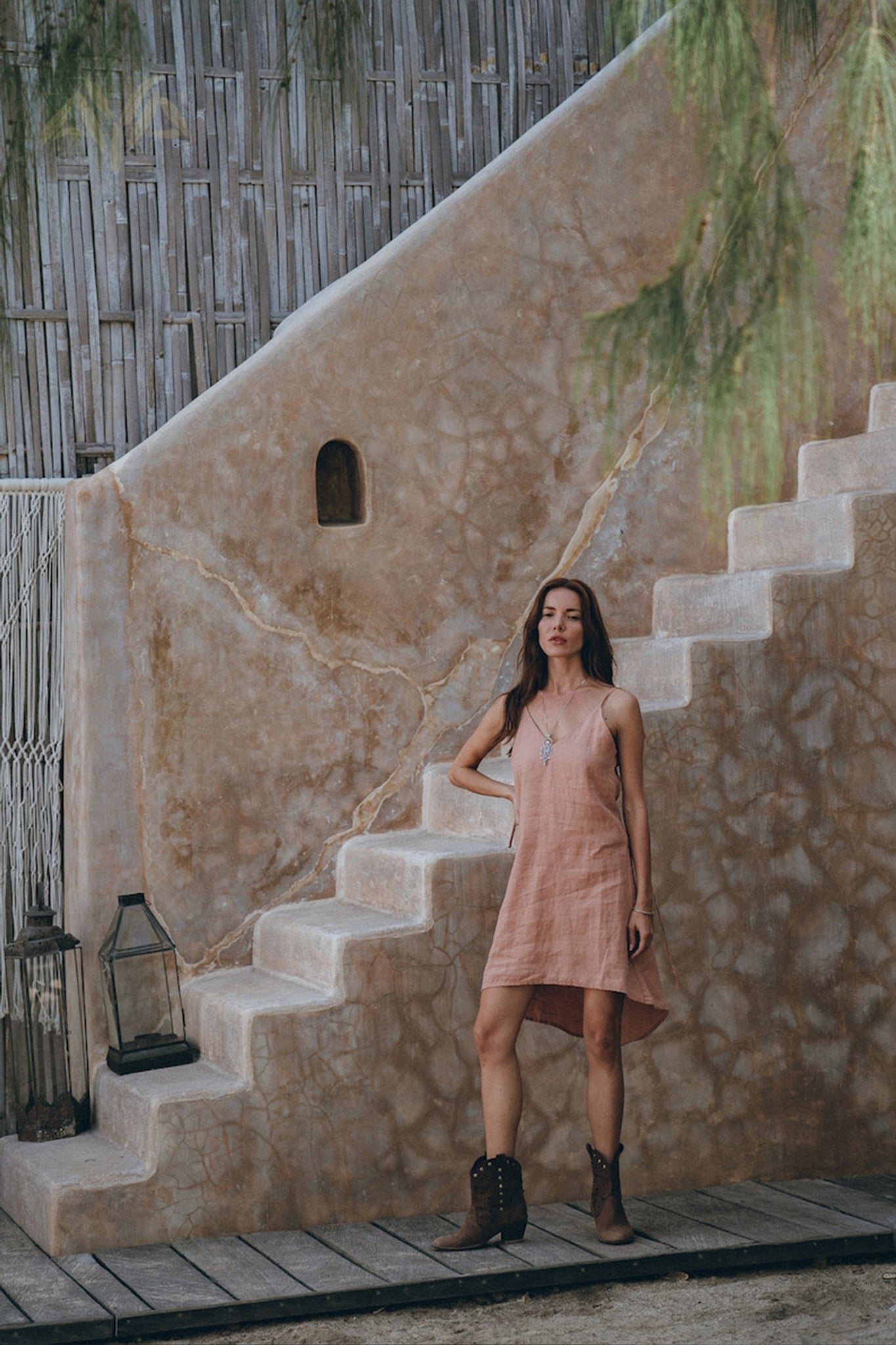 A woman in a sleeveless Pink Linen Bohemian Slip Midi Dress by AYA Sacred Wear and brown boots stands on wooden steps against a rustic stone wall. A lantern is placed nearby, with hanging greenery partially in view.