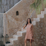 A woman in a sleeveless Pink Linen Bohemian Slip Midi Dress by AYA Sacred Wear and brown boots stands on wooden steps against a rustic stone wall. A lantern is placed nearby, with hanging greenery partially in view.