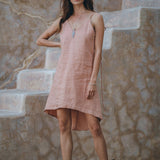 A woman wearing a delicate Pink Linen Bohemian Slip Midi Dress from AYA Sacred Wear and brown boots stands gracefully on textured stone steps. Her long hair flows freely, accented by a necklace, while the earthy tones of the wall behind add to the scene's natural charm.