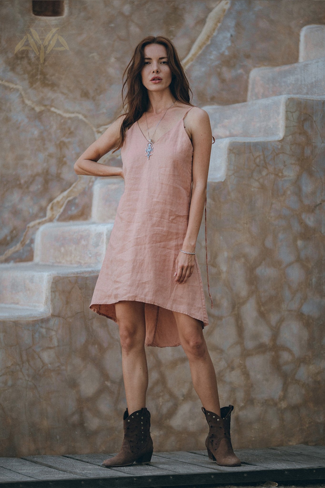 A woman wearing a delicate Pink Linen Bohemian Slip Midi Dress from AYA Sacred Wear and brown boots stands gracefully on textured stone steps. Her long hair flows freely, accented by a necklace, while the earthy tones of the wall behind add to the scene's natural charm.