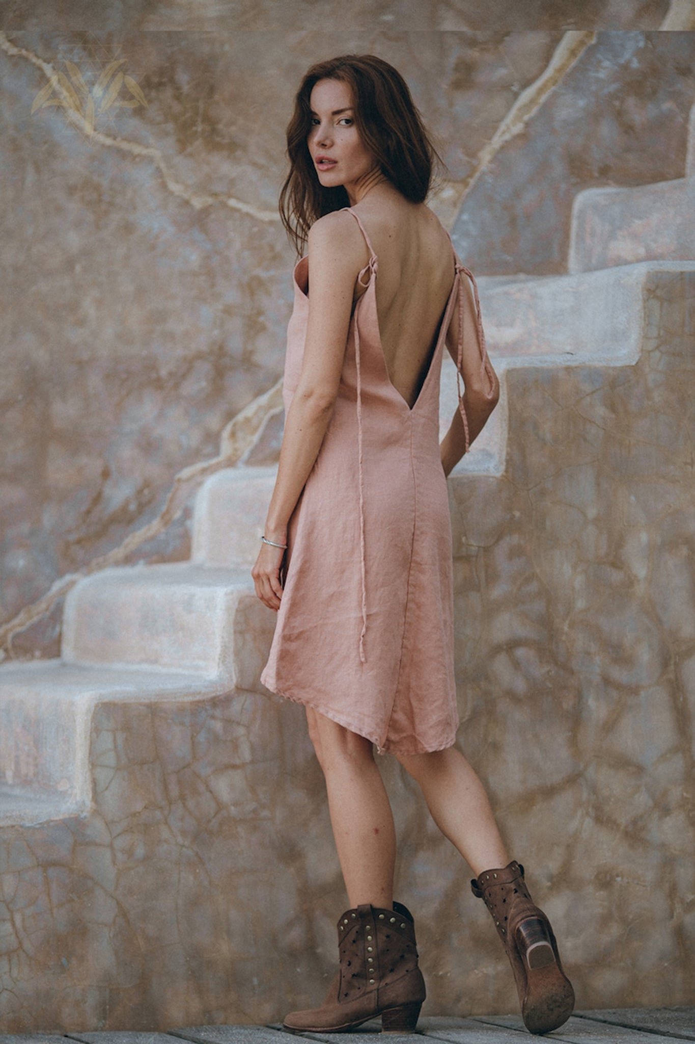 A woman with long hair stands on a stone staircase, wearing the Linen Boho Bridesmaid Dress by AYA Sacred Wear, a sleeveless, backless light pink bohemian midi dress made from organic linen. She pairs it with brown ankle boots and glances over her shoulder against a textured wall with natural tones.