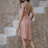A woman with long hair stands on a stone staircase, wearing the Linen Boho Bridesmaid Dress by AYA Sacred Wear, a sleeveless, backless light pink bohemian midi dress made from organic linen. She pairs it with brown ankle boots and glances over her shoulder against a textured wall with natural tones.