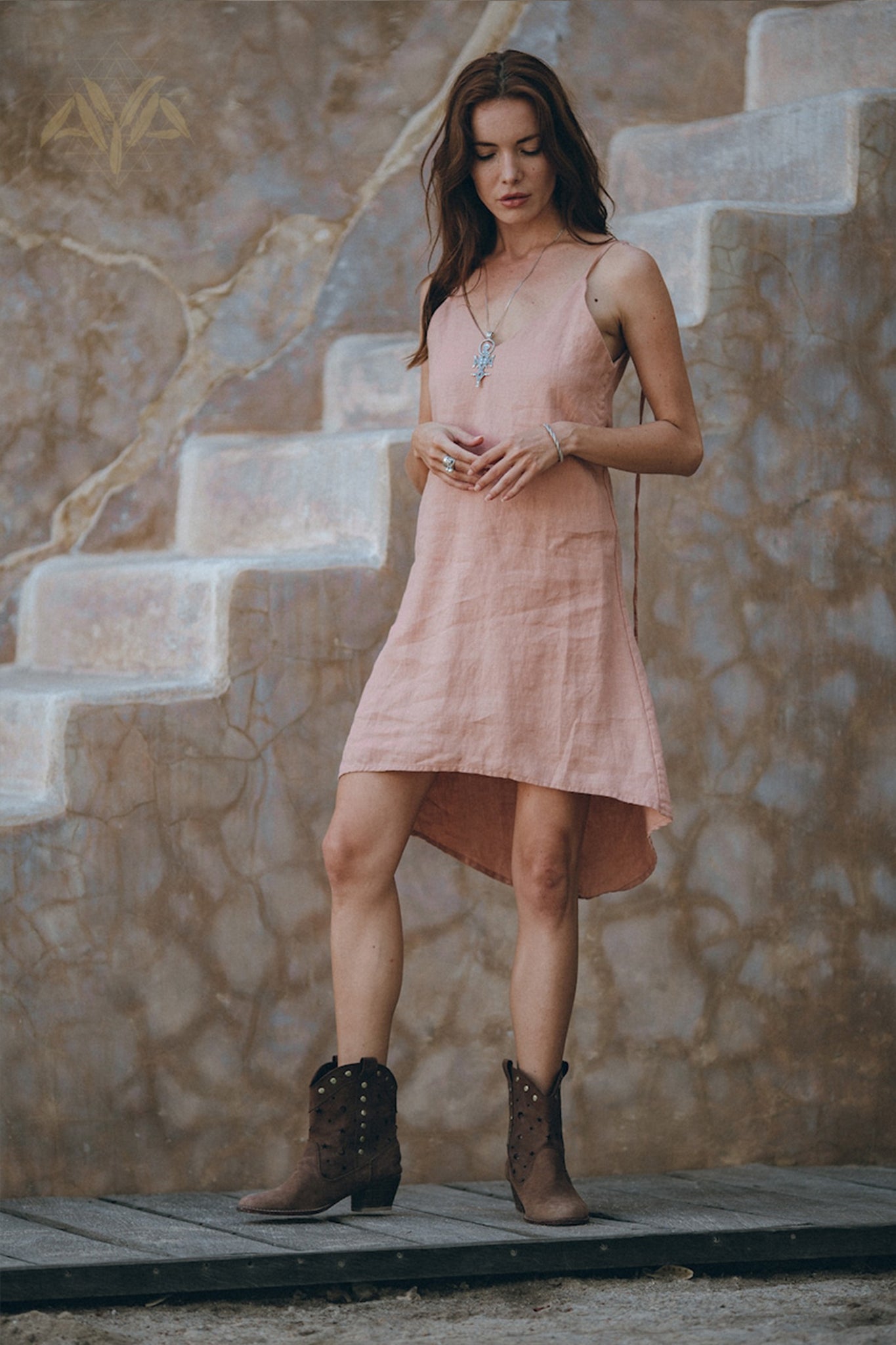 A woman wearing the AYA Sacred Wear Linen Boho Bridesmaid Dress in a pink hue pairs it with brown boots as she stands on a wooden floor. She gazes down, her hand gently touching a necklace. In the background, a rustic wall with faded stairs enhances the scene's earthy, vintage vibe with its sustainable ambiance.